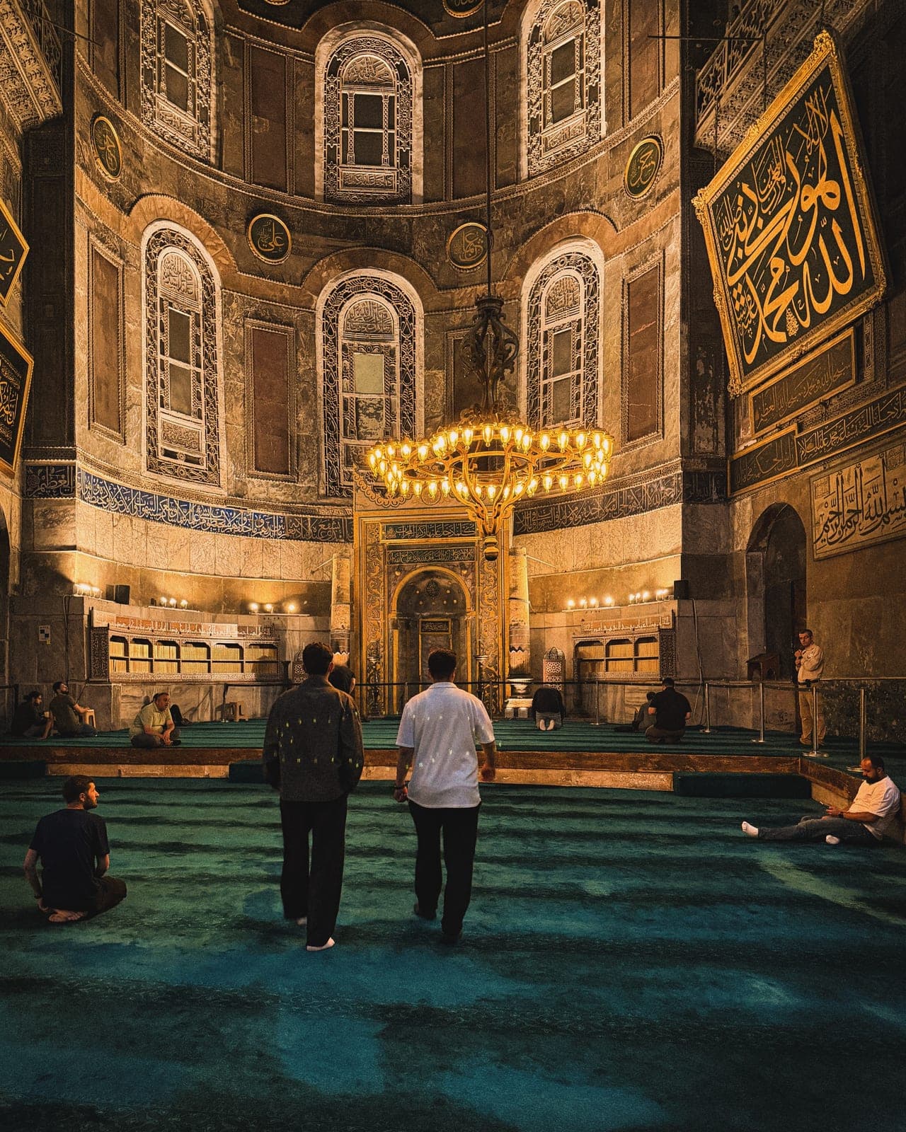 Worshippers inside a mosque arcade, lit by hanging lamps